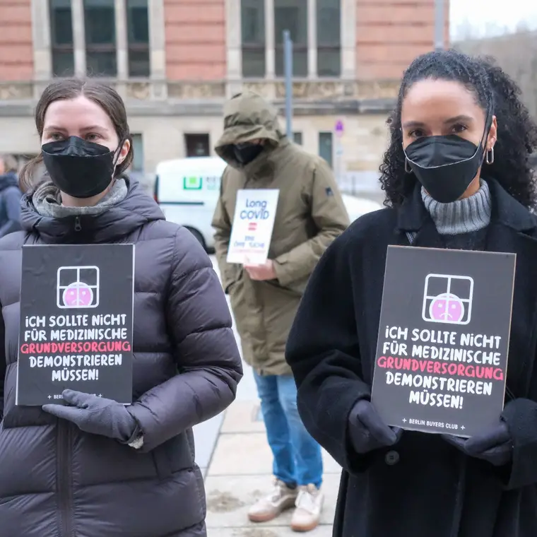 Two people in masks holding signs: I shouldn’t have to protest for basic medical care